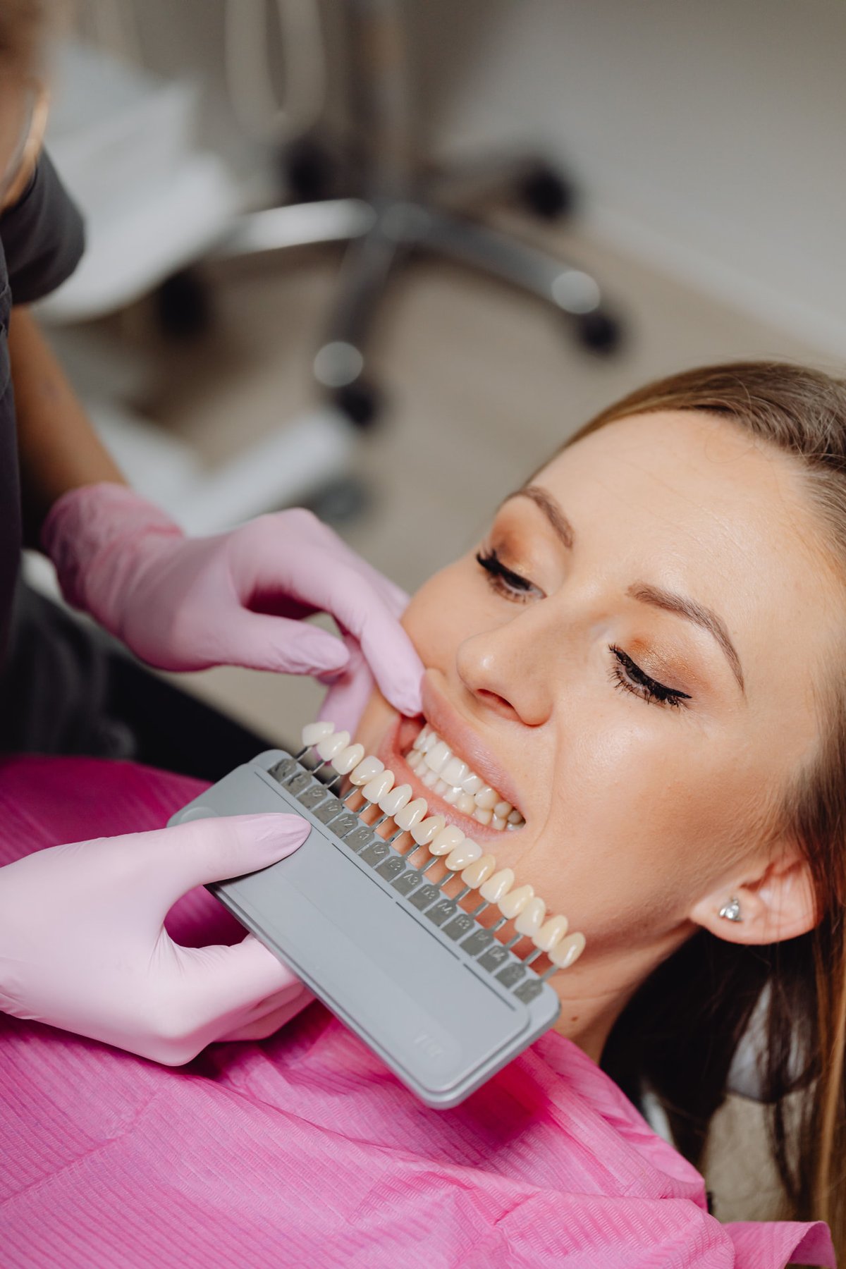 Patient reclined in a dental chair while a dentist compares tooth shades with a shade guide against her smile